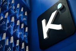 Illuminated Kimi logo mounted on a blue wall beside rows of branded tote bags at a Moonshot AI event